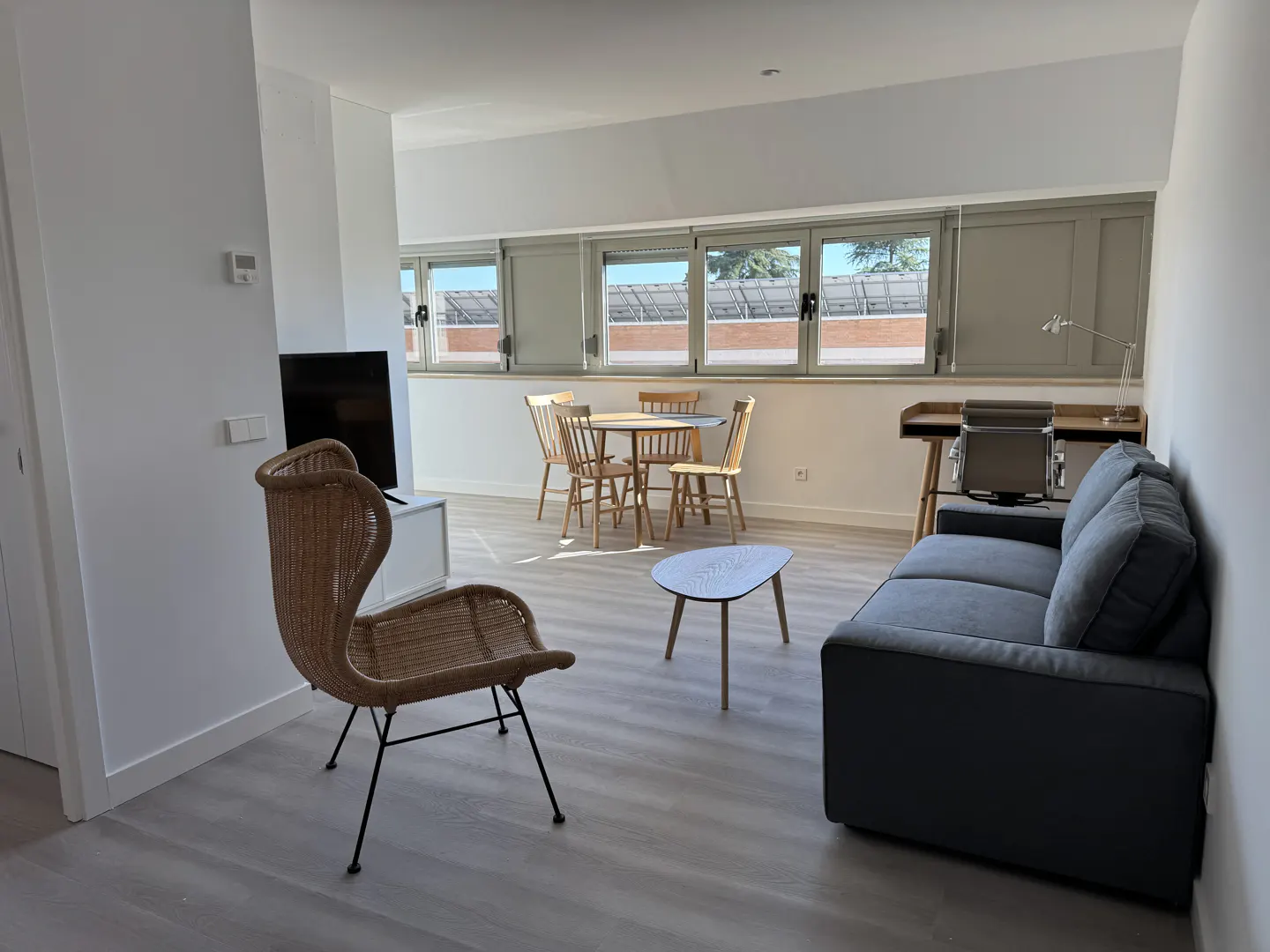 Bright, open living space with a gray sofa, wicker chair, and round table with four chairs. A desk sits by the window.