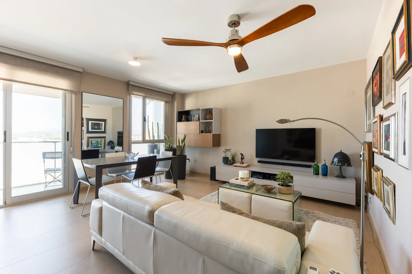 A bright living room with a white sofa, dining table, TV, and a wooden ceiling fan. Large windows offer a view of the outside.