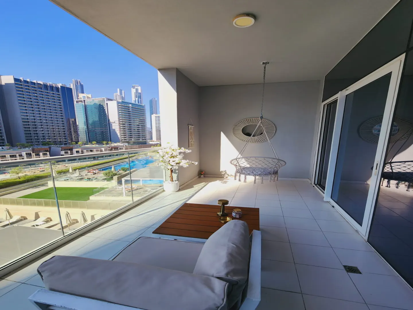 A modern balcony with a white couch, wooden table, and hanging chair overlooking a city skyline.