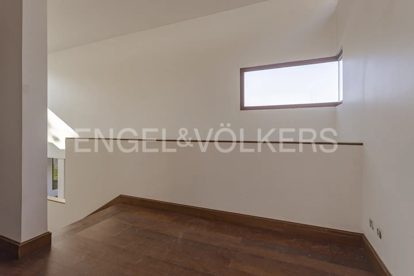 Interior view of a modern home featuring dark wood floors, white walls, and a small window. The Engel & Völkers logo is visible on the wall.