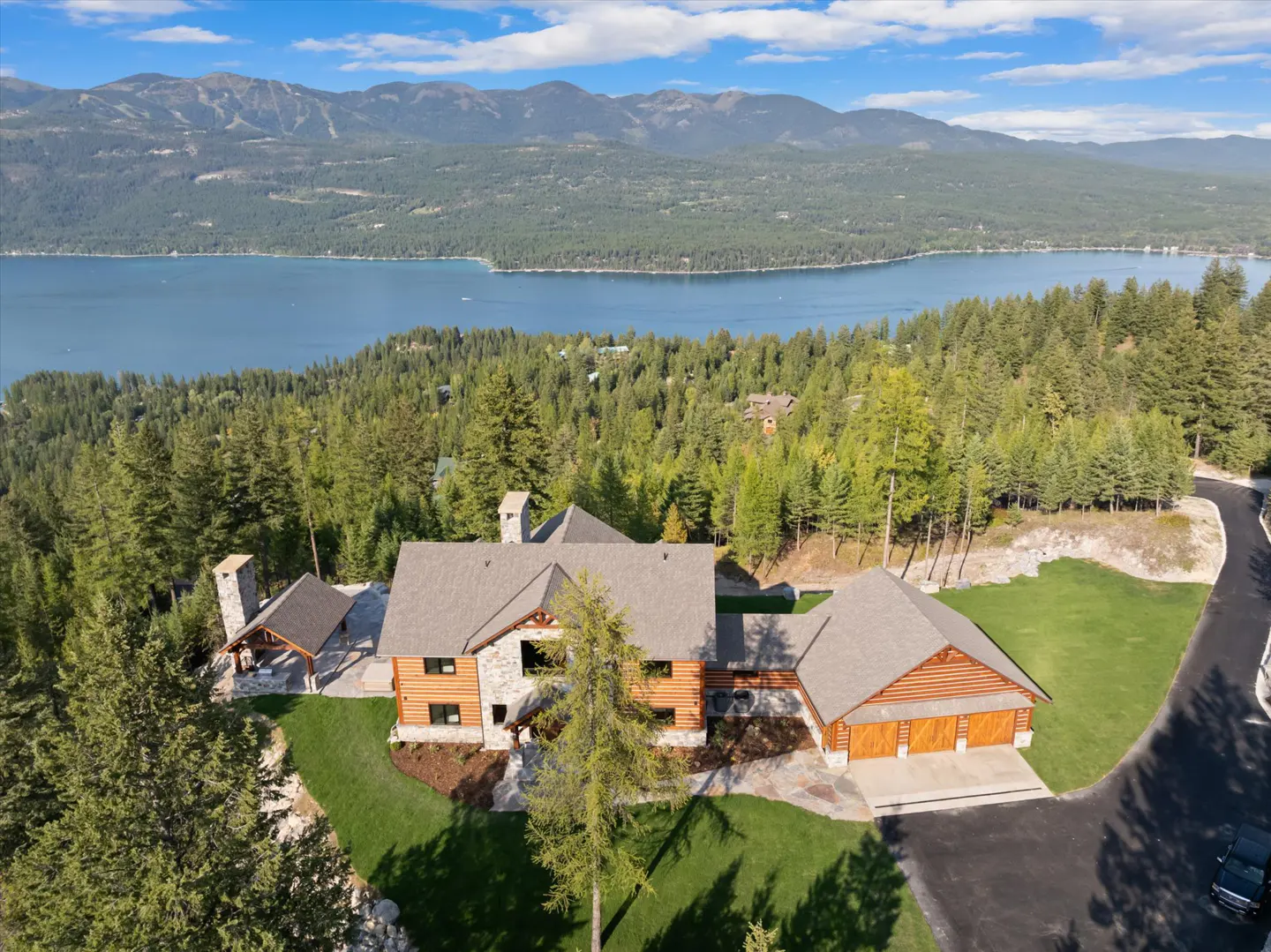 Aerial view of a log cabin home with a lake and mountains in the background. The house has a stone chimney and a two-car garage.