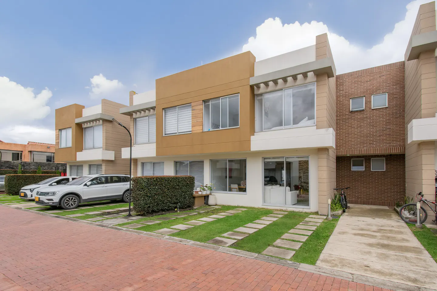 Modern townhouses with tan and white exteriors, cars parked in front, and brick paved street.