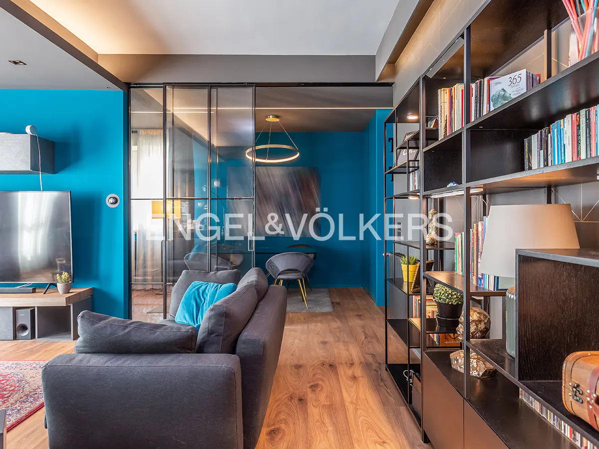 Living room with blue walls, a gray sofa with a blue pillow, and a black bookshelf filled with books.