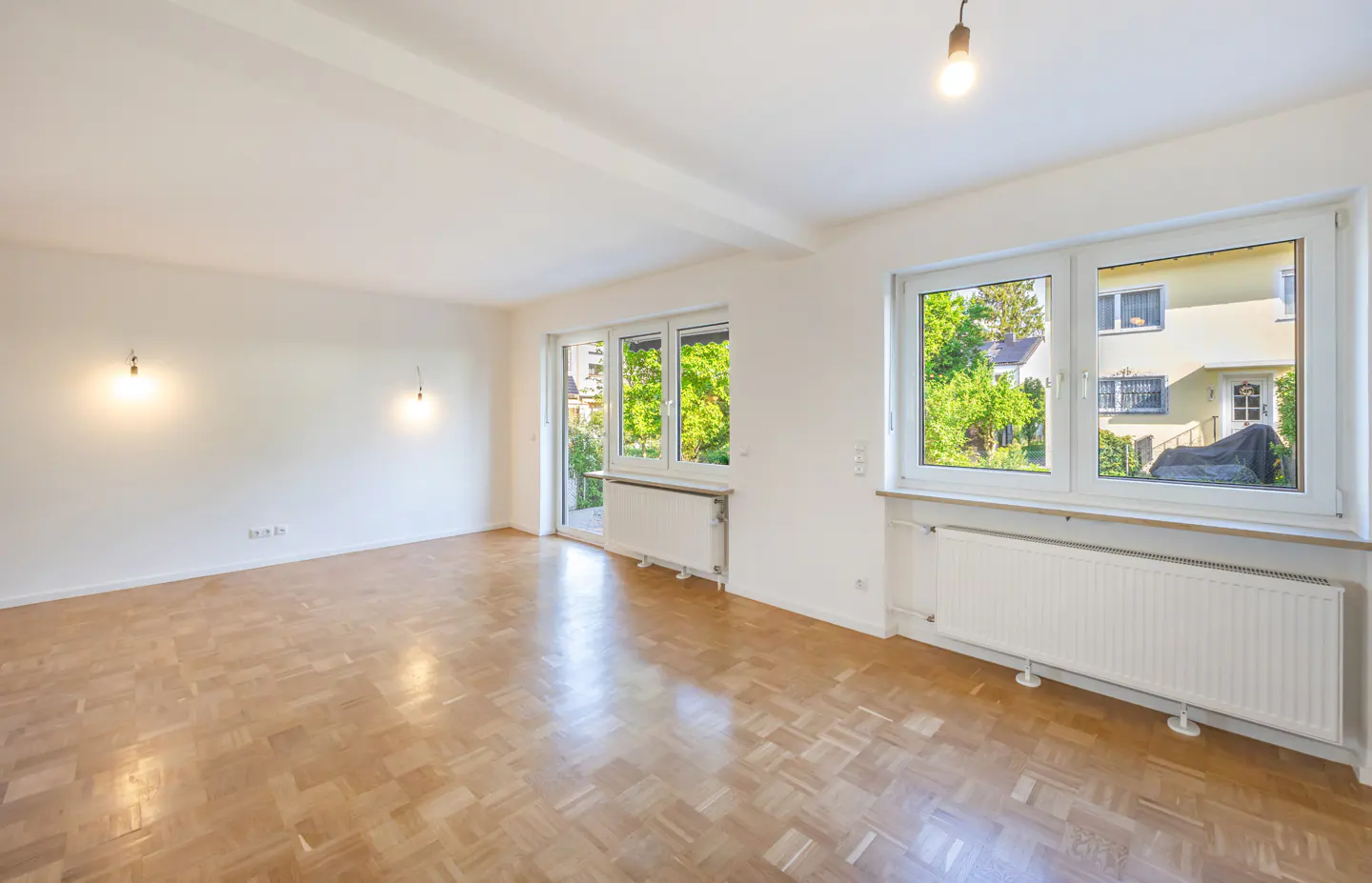 Bright, empty room with parquet floors, white walls, and large windows showing a green yard and neighboring house.