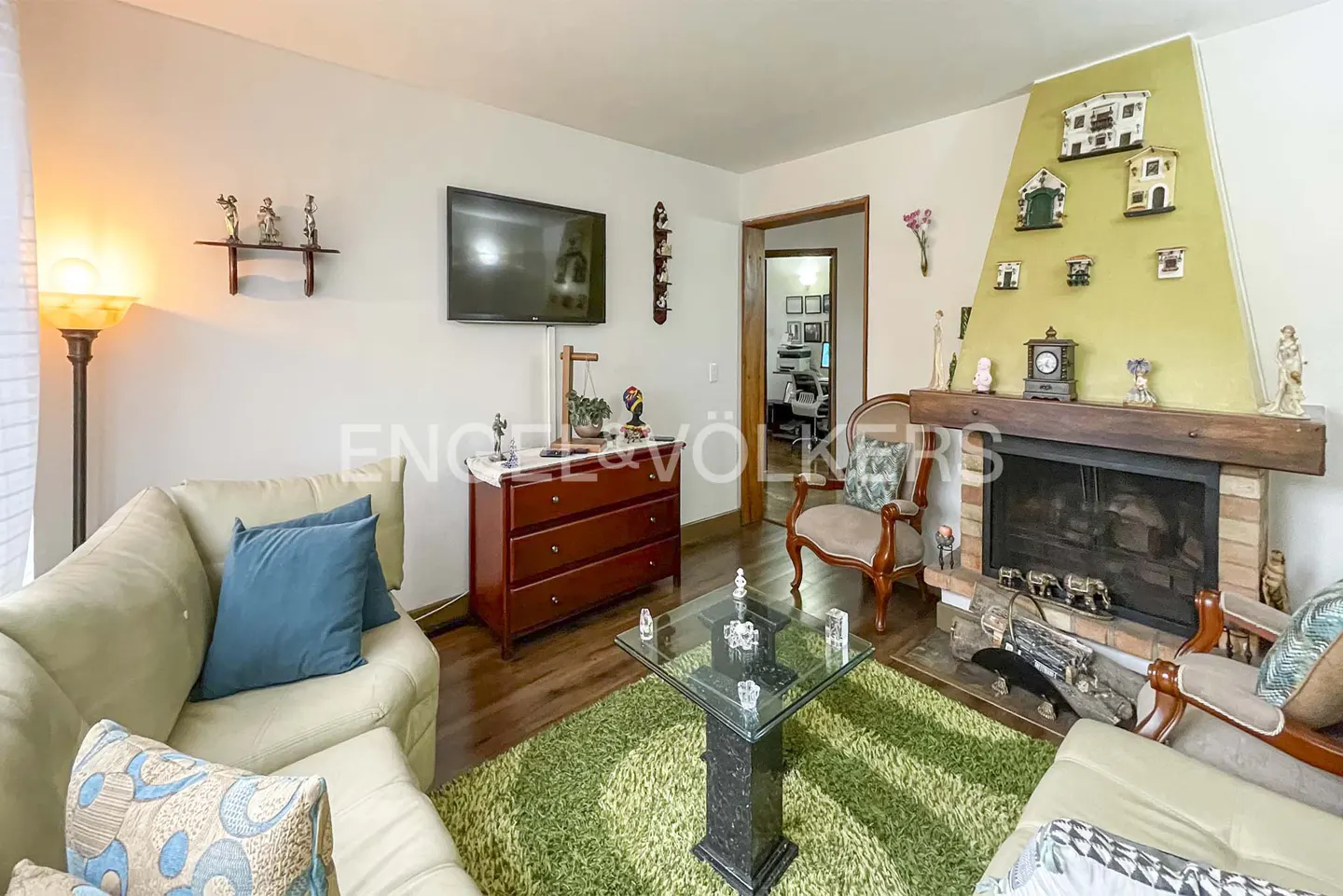 A cozy living room with a fireplace, sofas, a glass table, and a TV. The walls are white, and the floor is wooden.