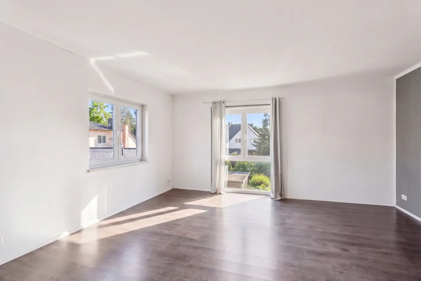 Empty room with white walls, dark wood floors, and two windows looking out onto a residential neighborhood.
