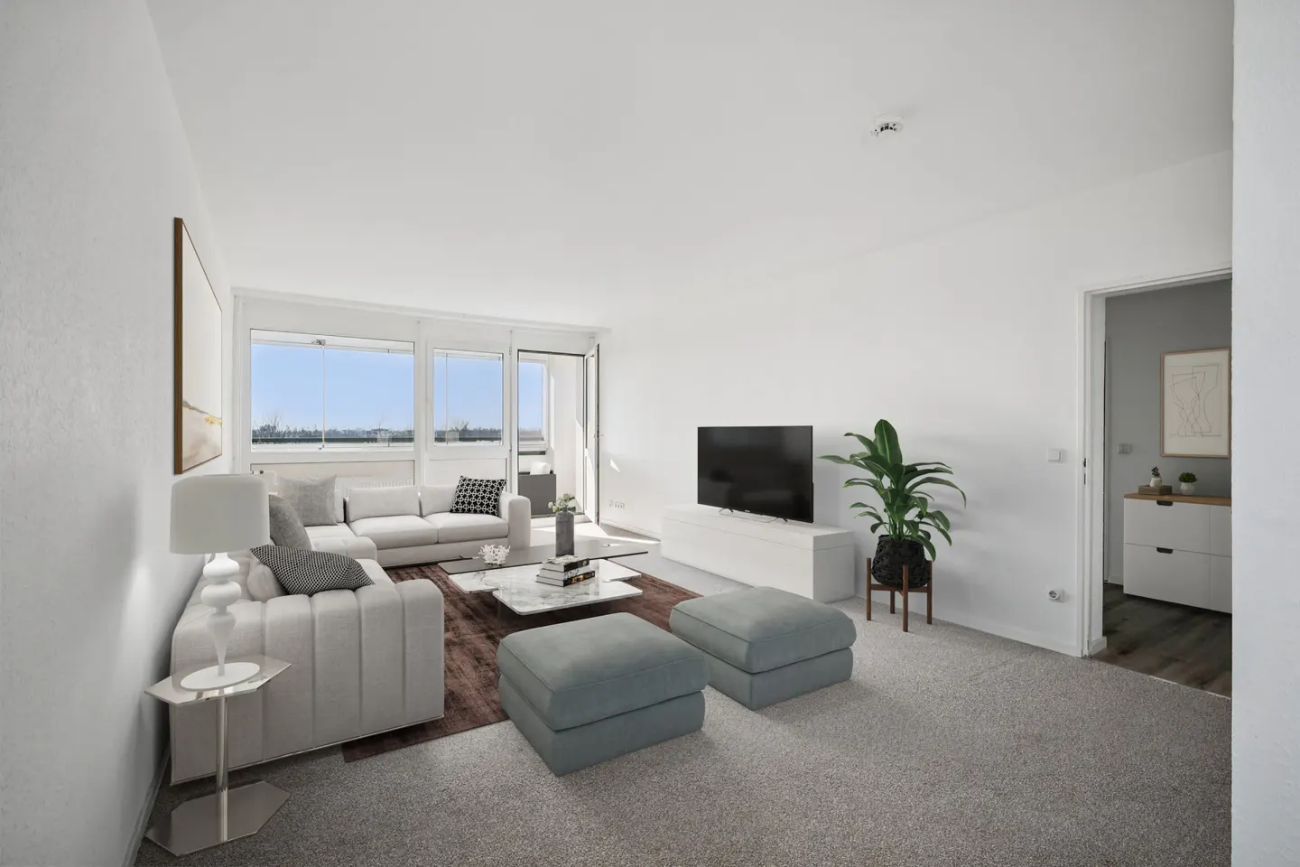Bright living room with white walls, gray carpet, and large windows. A white sofa, ottomans, TV, and plant create a modern, airy space.