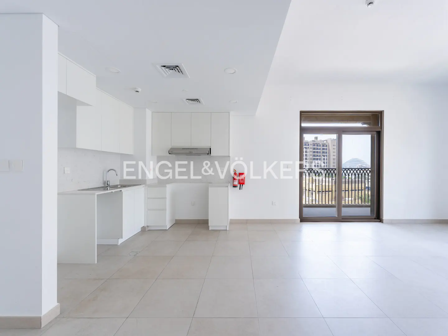 Bright, empty apartment with white walls, tile floors, and a white kitchen. Sliding glass doors lead to a balcony with a view.