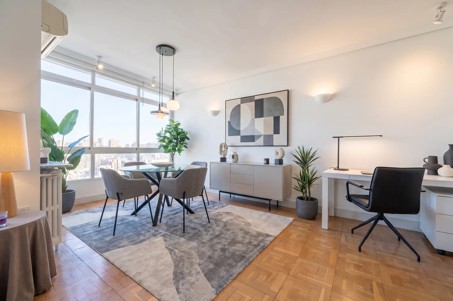 Bright, modern apartment interior with a glass dining table, gray chairs, a desk, and a large window overlooking a city.