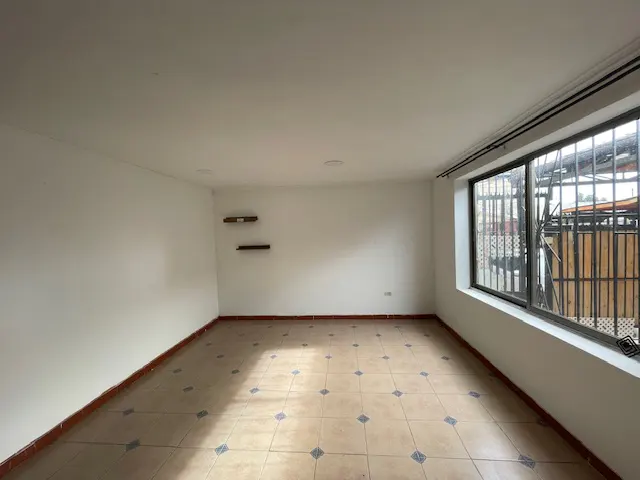 Empty room with white walls, tile floor, and a large window with bars. Two small shelves are on the left wall.