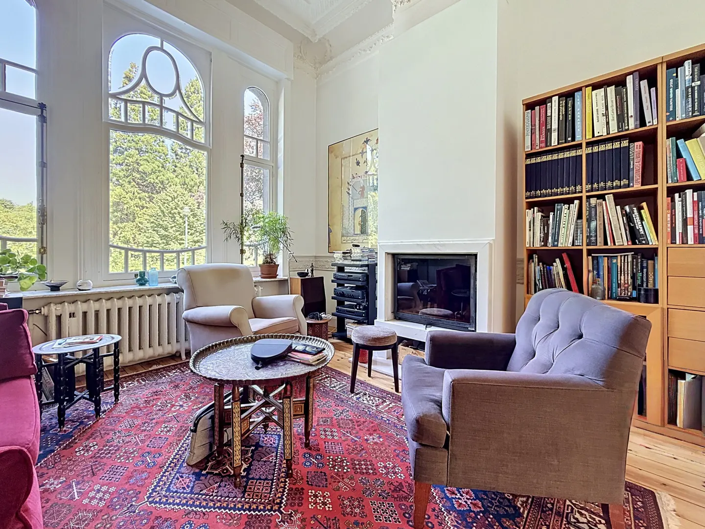 Bright living room with large windows, red rug, beige and purple armchairs, fireplace, and a full bookshelf.