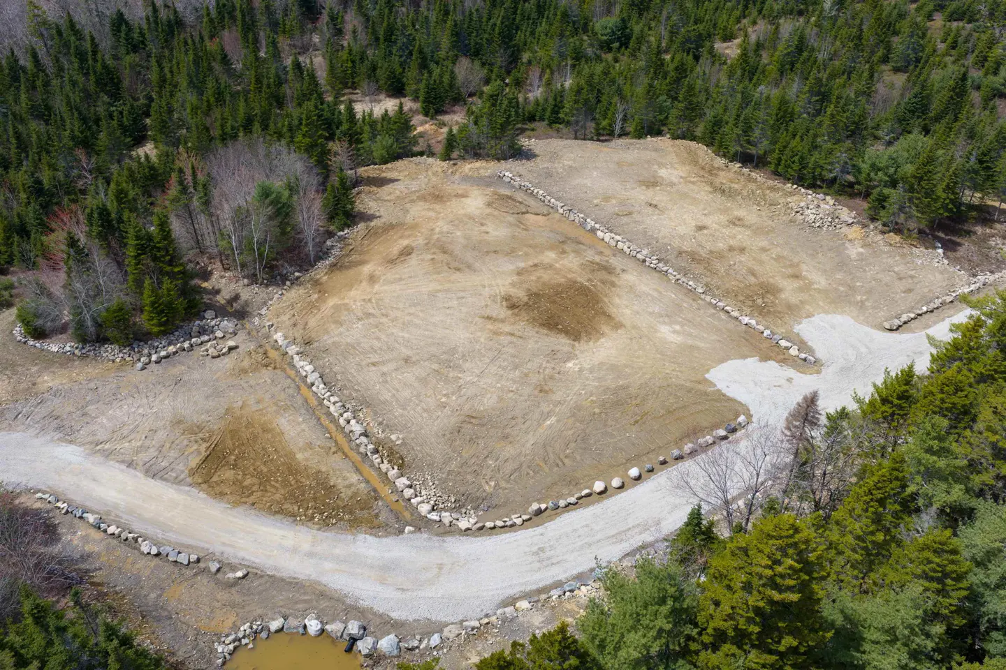 Aerial view of cleared land, bordered by rocks, surrounded by green trees. A gravel road cuts through the land.