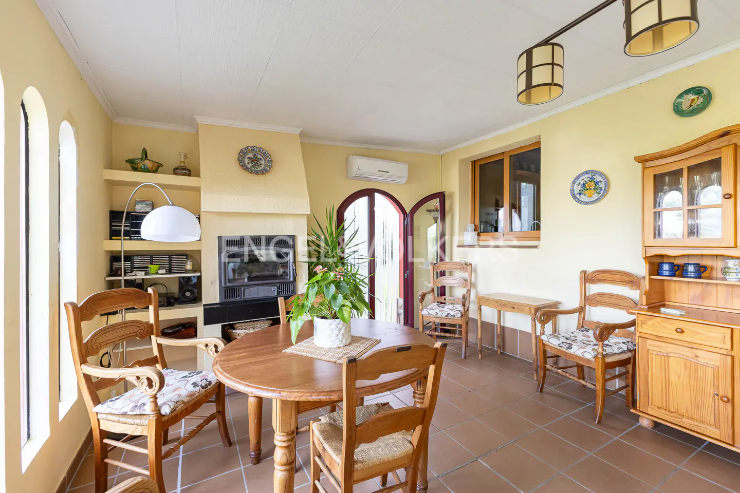 A sunroom with yellow walls, a round wooden table, and chairs. A fireplace, shelves, and a wooden hutch add charm. Tiled floors and natural light complete the scene.