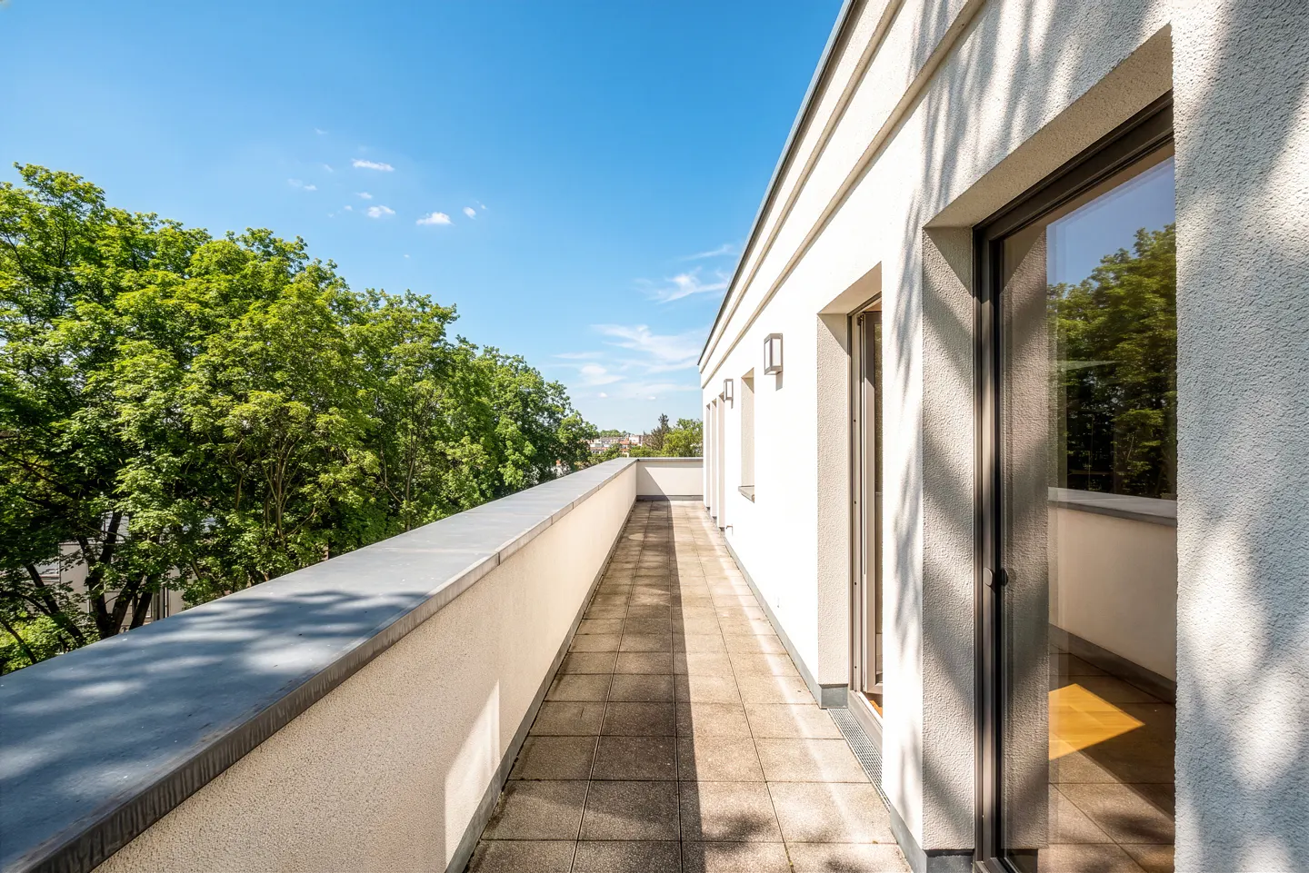 Long, narrow balcony with gray tiled floor and white walls, overlooking green trees under a blue sky.