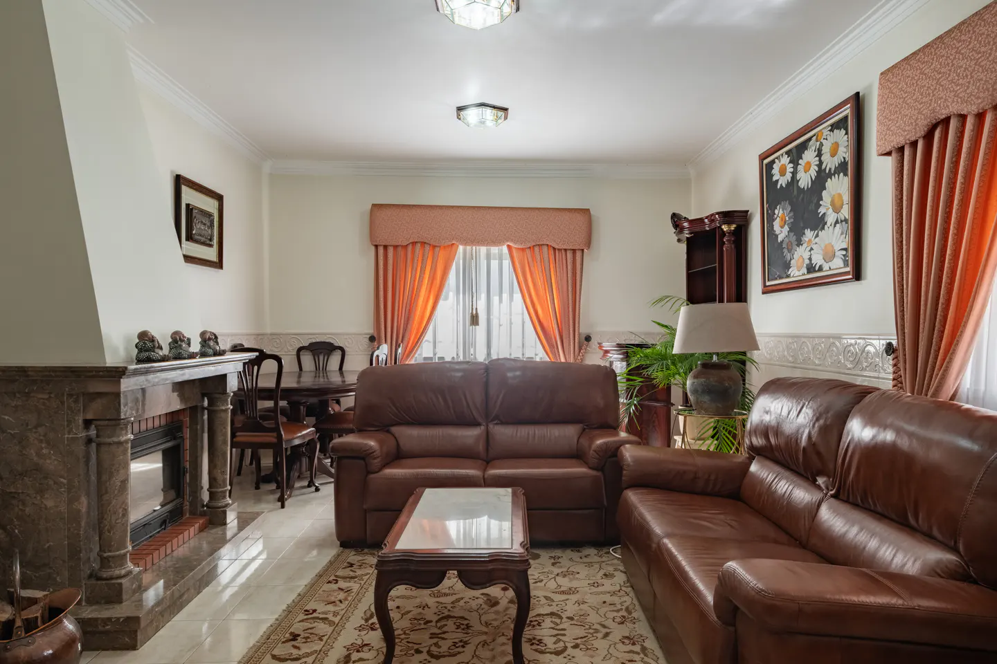 Living room with brown leather sofas, marble fireplace, dining table, and orange curtains. A patterned rug covers the tile floor.