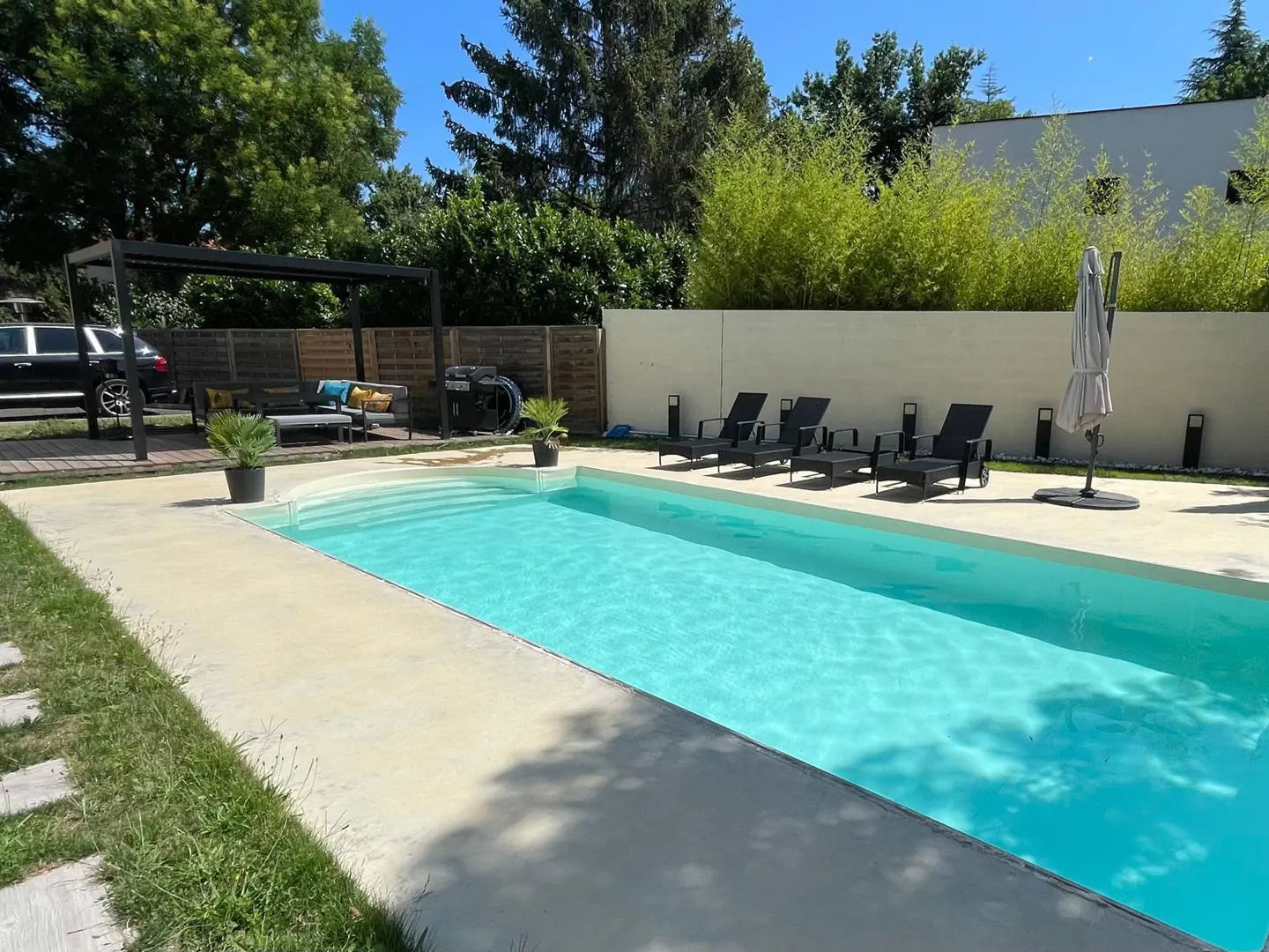 A backyard swimming pool with lounge chairs, a pergola, and green trees under a blue sky.