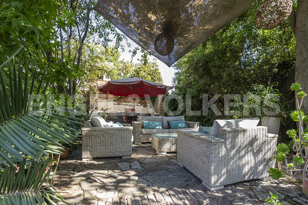 Outdoor patio with wicker furniture, blue cushions, and stone flooring. A red umbrella and lush greenery provide shade.