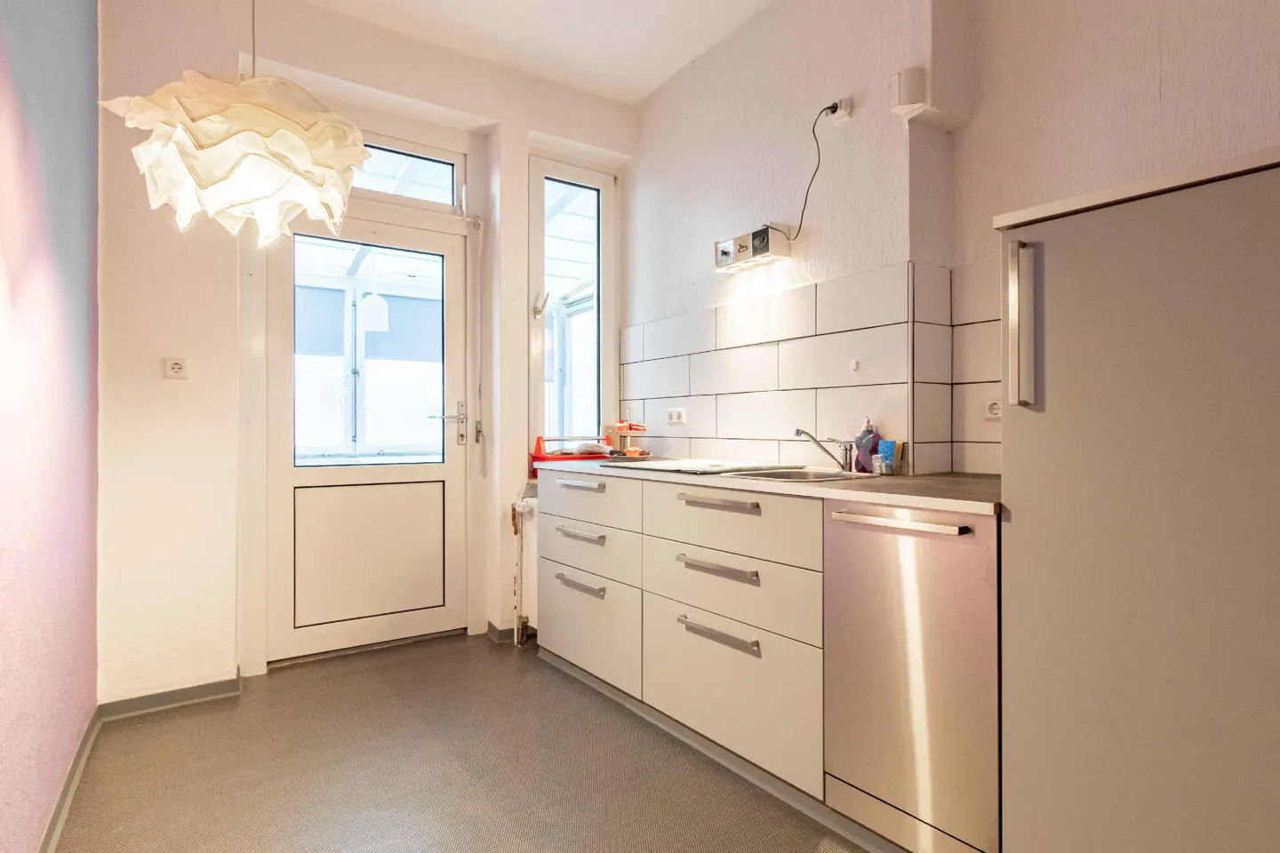 Bright kitchen with white cabinets, stainless steel dishwasher, and a door leading outside. A modern white lamp hangs from the ceiling.