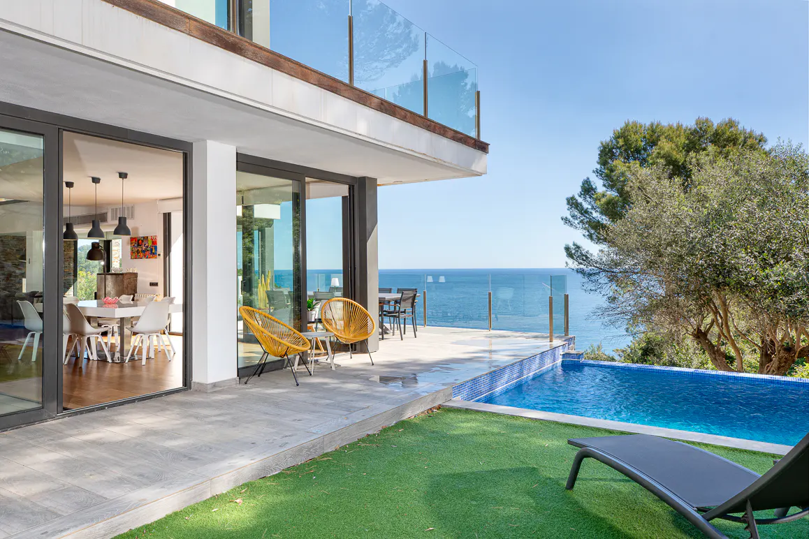 Modern white house with a pool overlooking the ocean. Yellow chairs sit on the patio. Dining room visible through sliding glass doors.