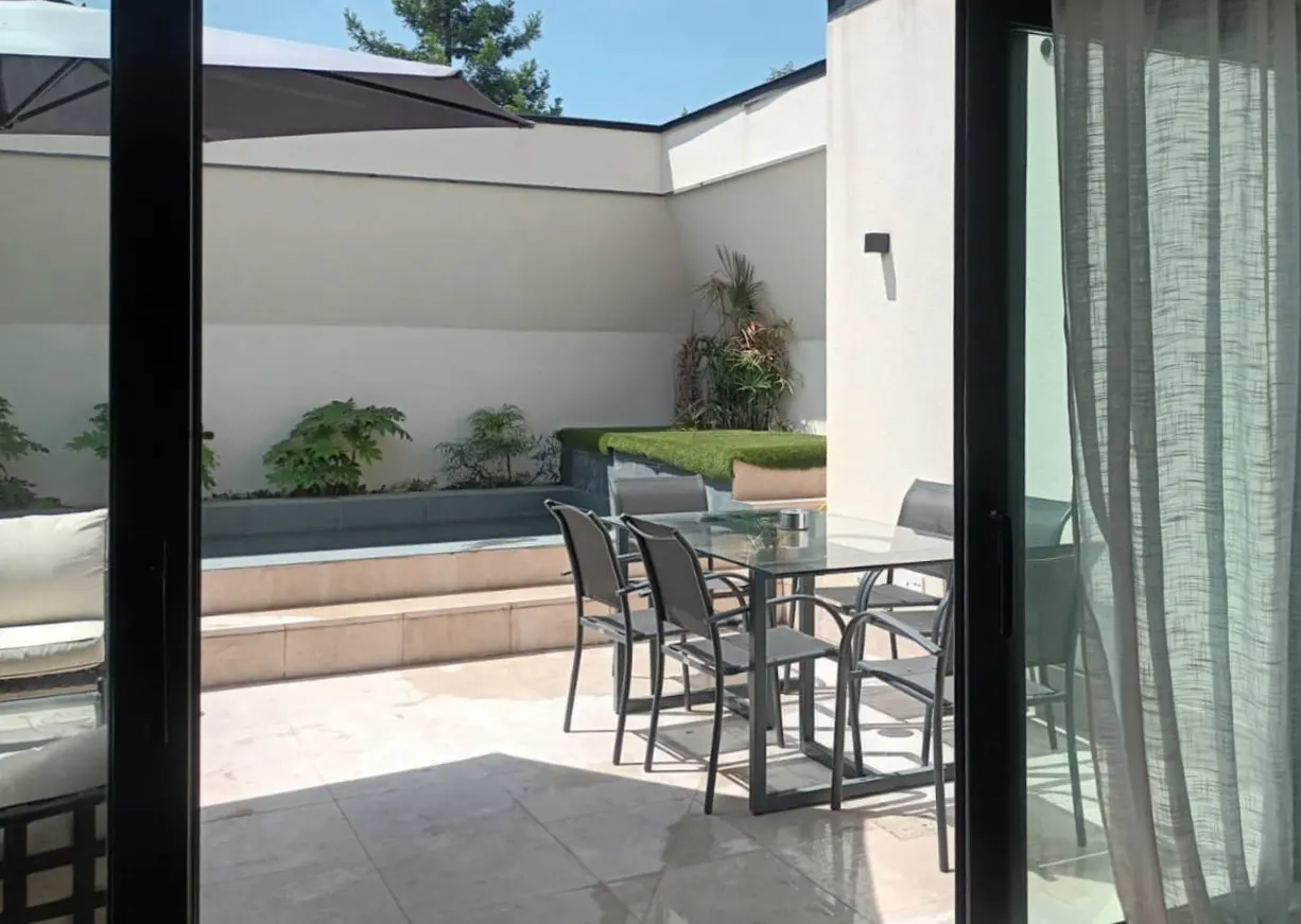 View from inside to a patio with a glass table, six chairs, and a small pool with plants. The patio has light-colored tiles and white walls.