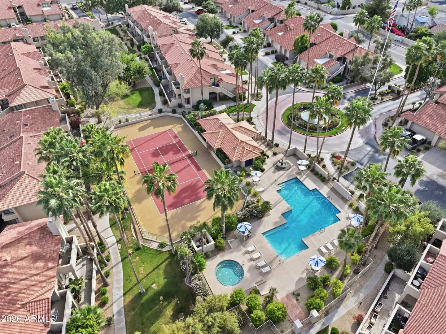 Aerial view of a community with a pool, tennis court, palm trees, and red-tiled roofs.