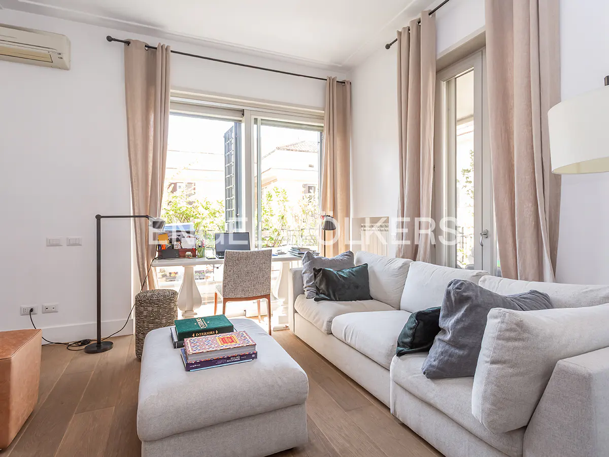 Bright living room with a white sectional sofa, matching ottoman, and a desk by a window with beige curtains.