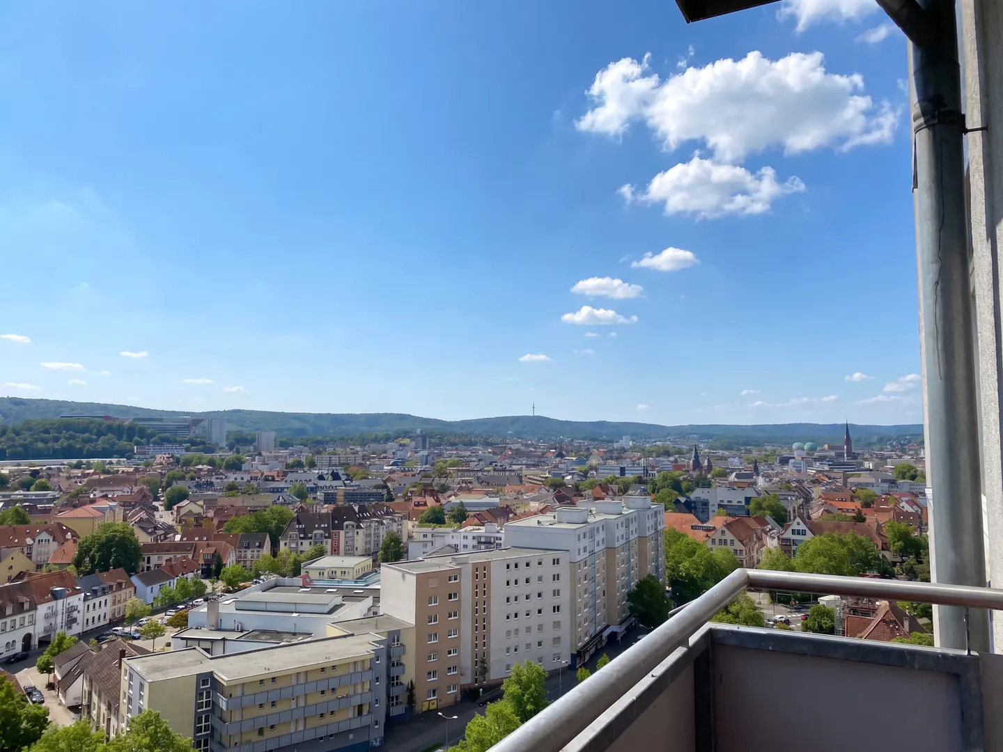 View from a balcony overlooking a city with red-roofed buildings, green trees, and a blue sky with white clouds.