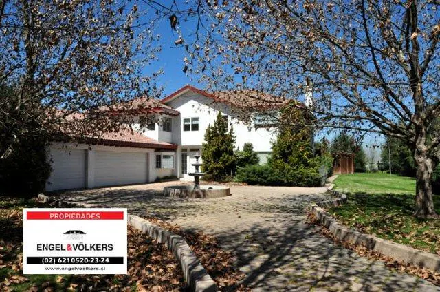 A two-story white house with a red tile roof, a fountain in the front yard, and trees with bare branches. An Engel & Völkers sign is in the foreground.