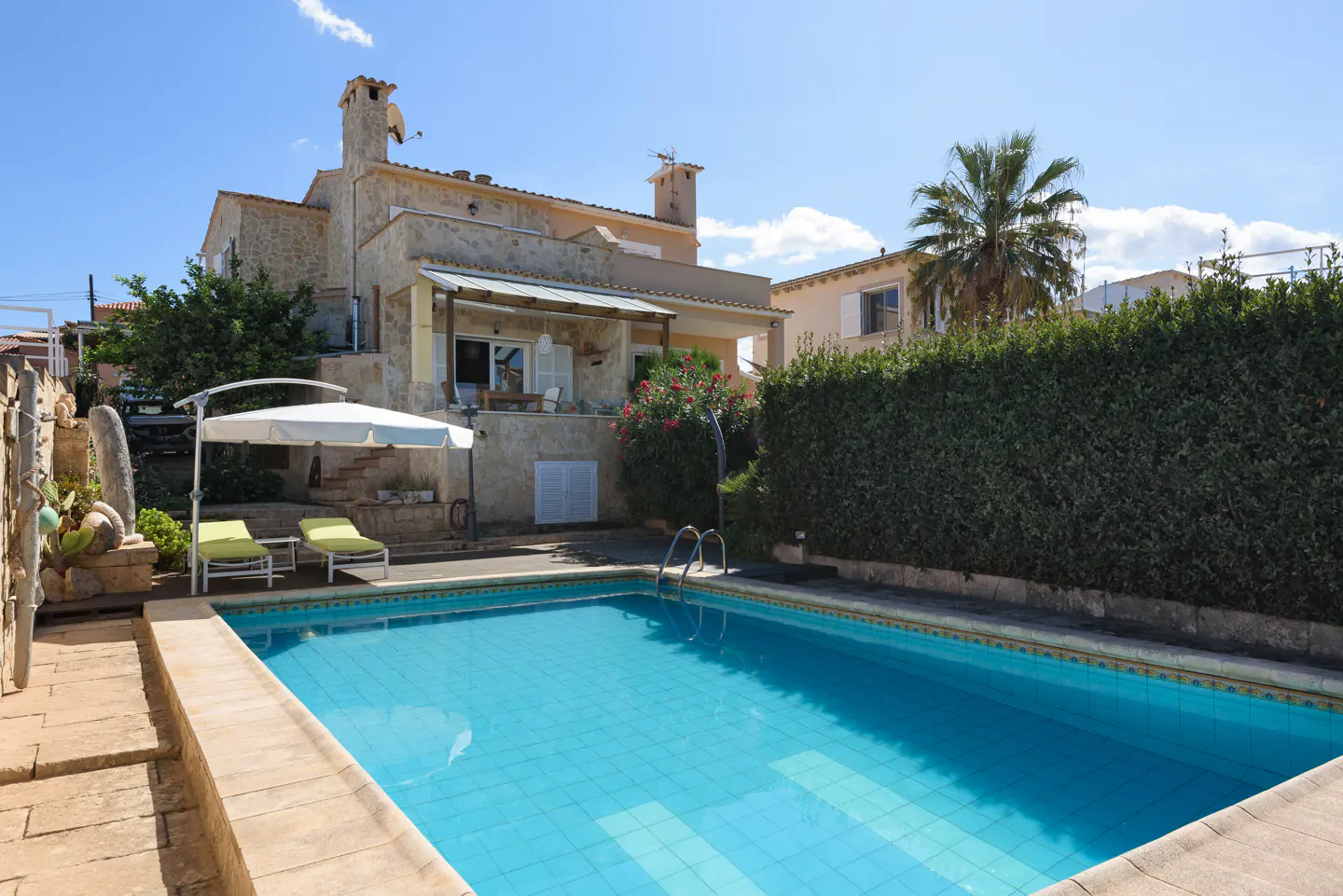 A backyard with a blue tiled pool, lounge chairs, and a stone house under a sunny sky.