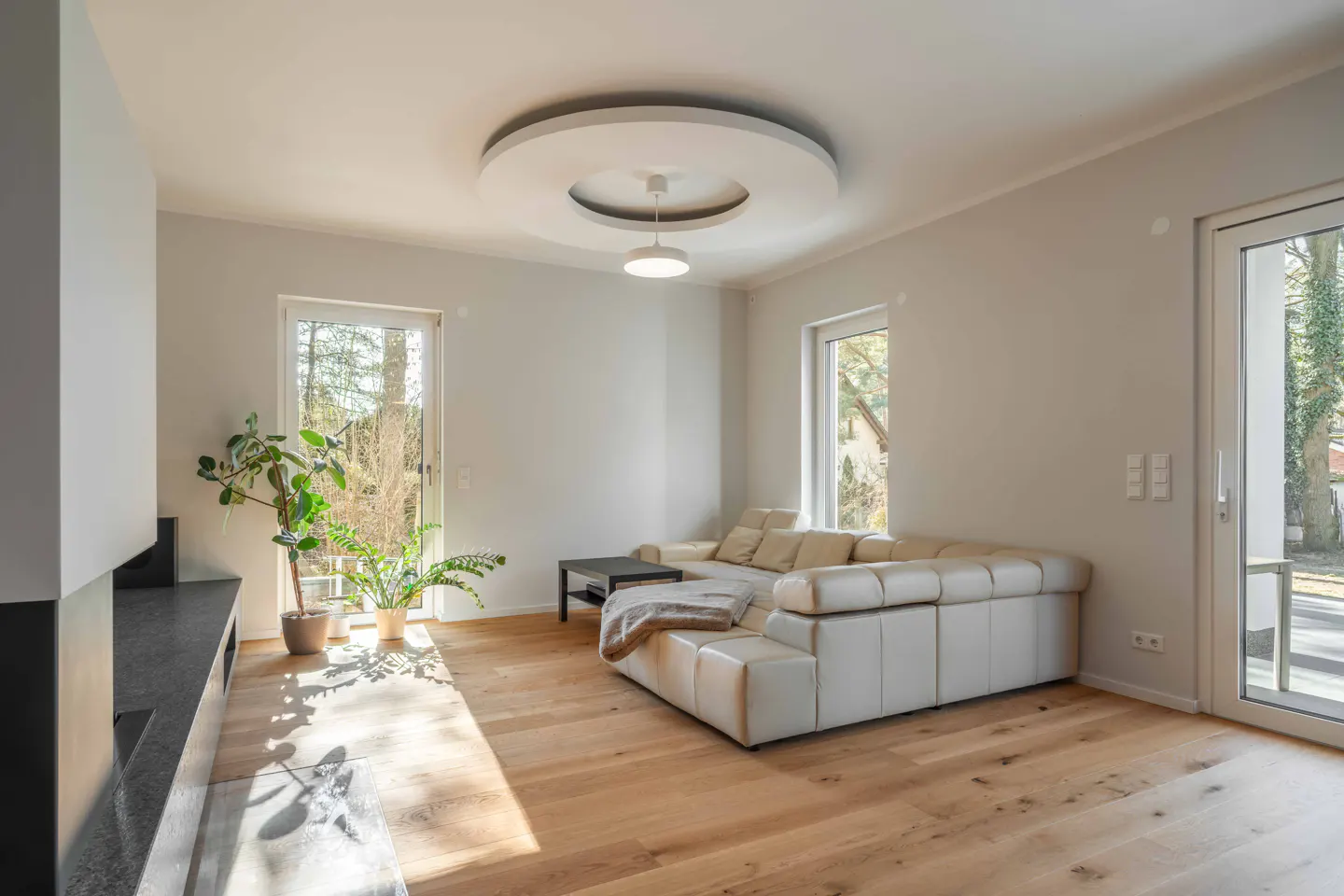 Bright living room with hardwood floors, white walls, and a white sectional sofa. Windows and a glass door let in natural light.
