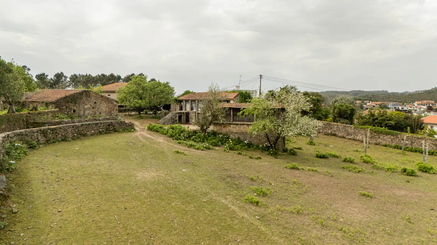 Exterior view of stone houses with red tile roofs, surrounded by green grass and trees under a cloudy sky.