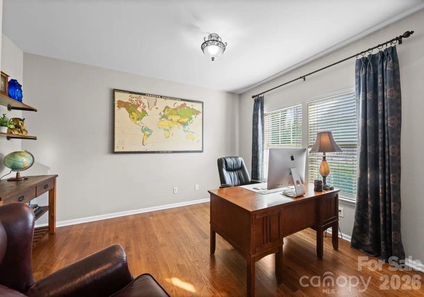 A home office with a wooden desk, computer, and a world map on the wall. There is a leather chair and dark curtains.