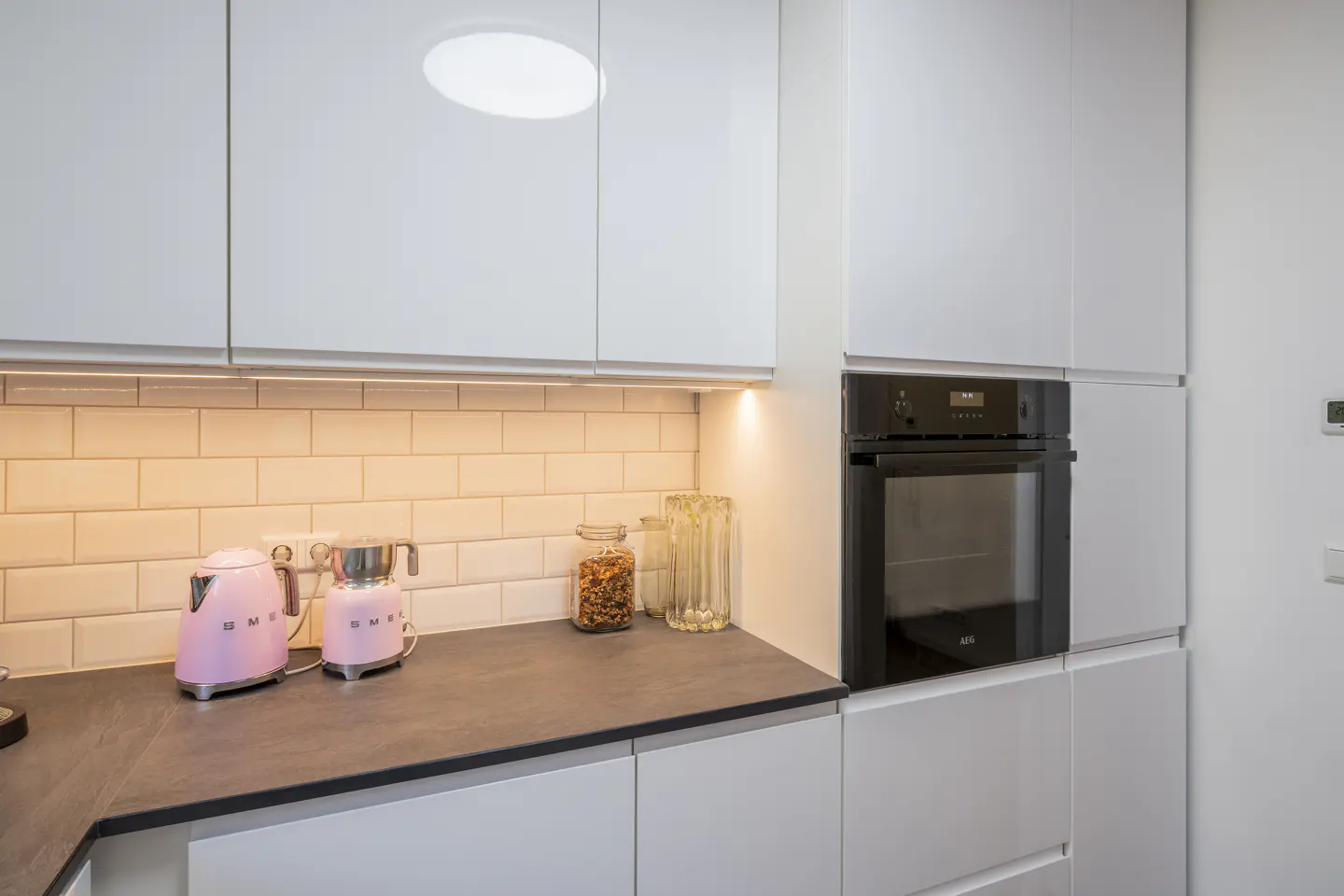 A modern kitchen with white cabinets, a black oven, and a gray countertop. Two pink Smeg appliances sit on the counter.