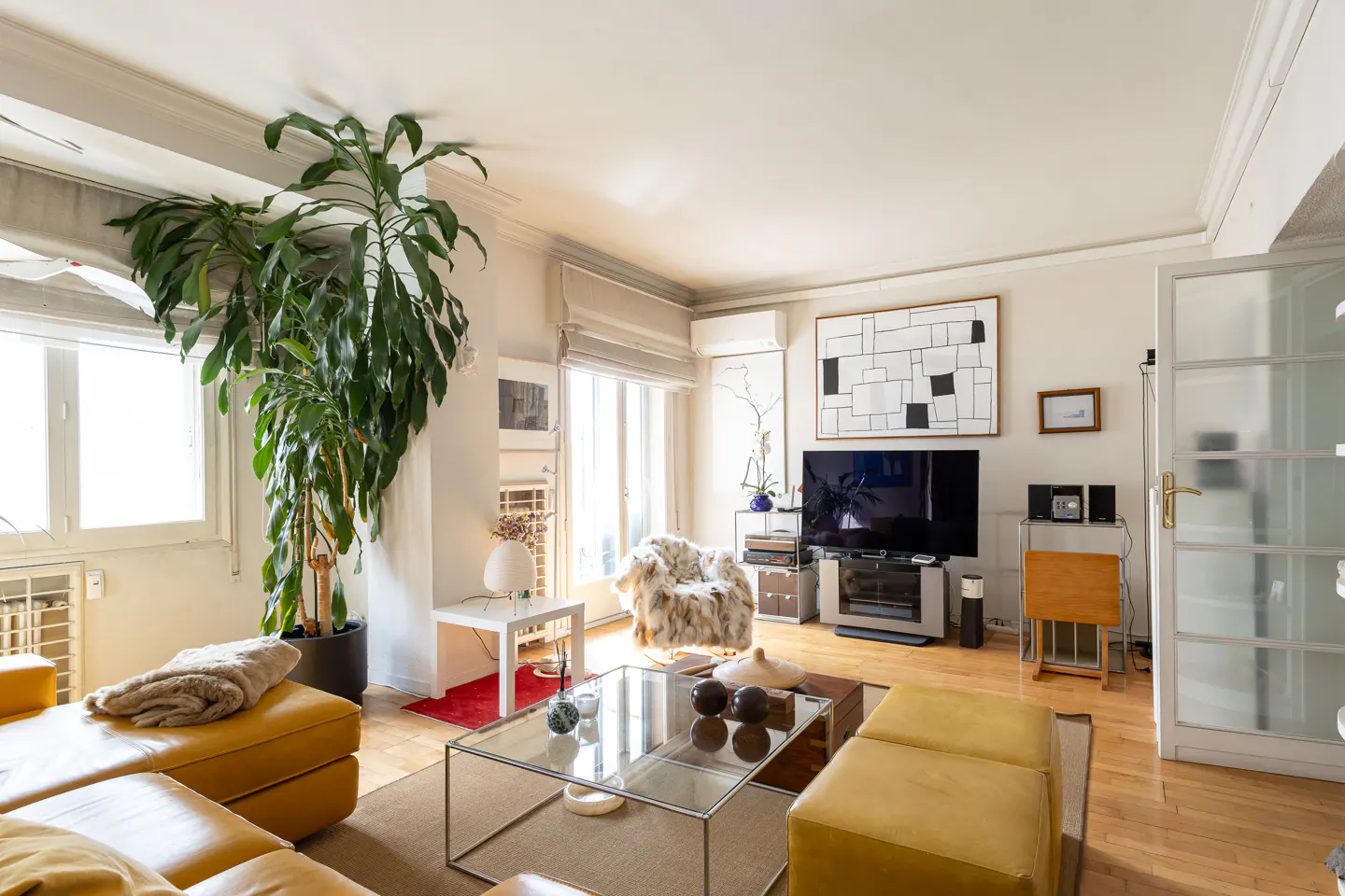 Bright living room with yellow sofa, glass table, and large potted plant by the window. TV and art on the wall.