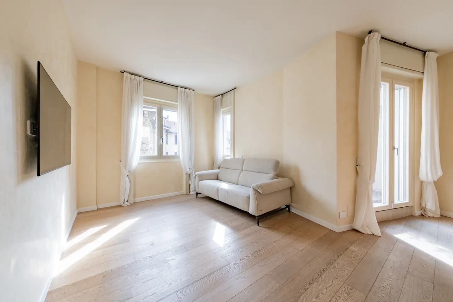 Bright living room with wood floors, cream walls, and a light beige sofa. A flat-screen TV hangs on the wall, and windows have sheer white curtains.