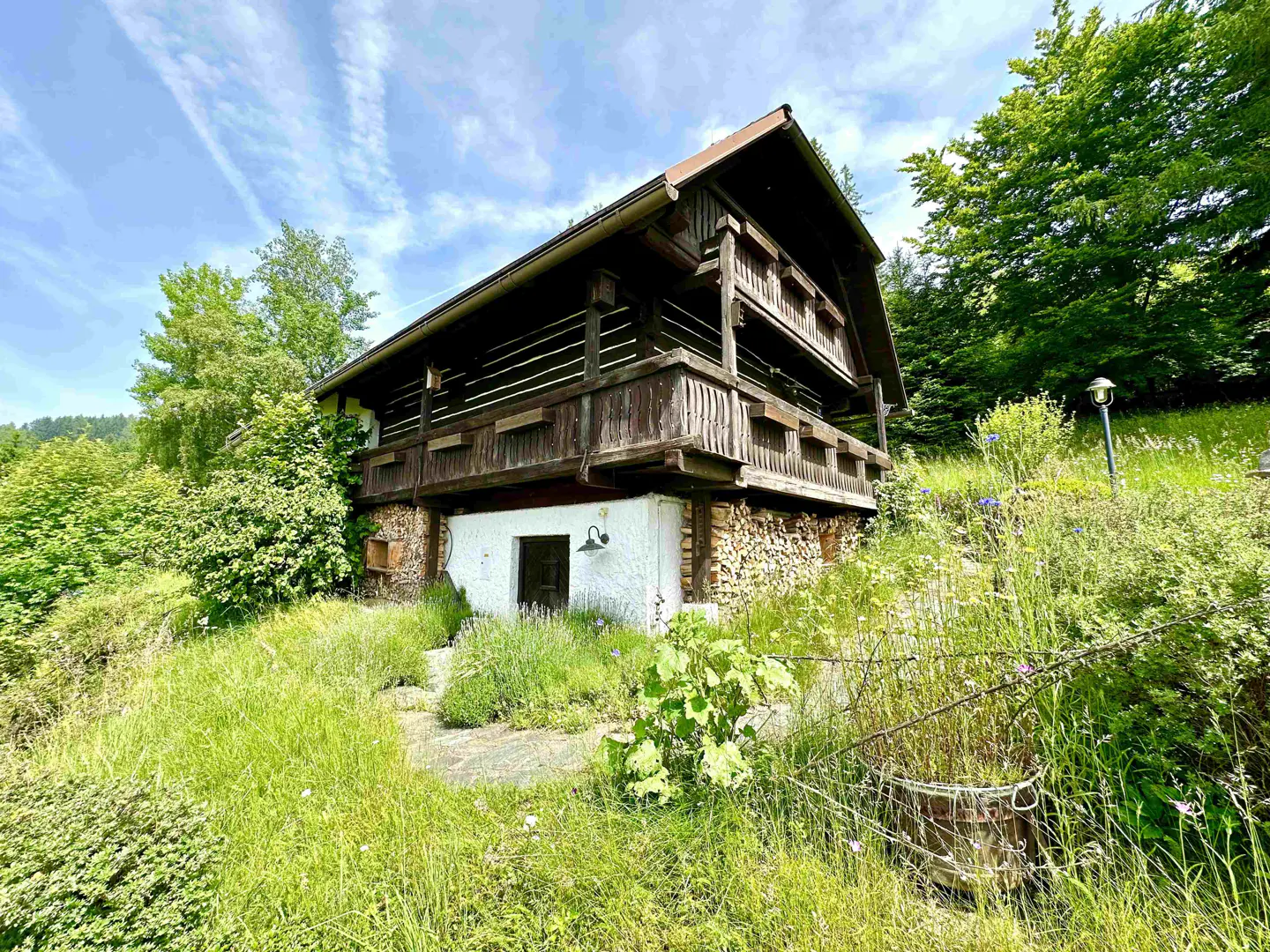 A two-story log cabin with balconies, a white stone foundation, and a woodpile sits on a grassy hill.