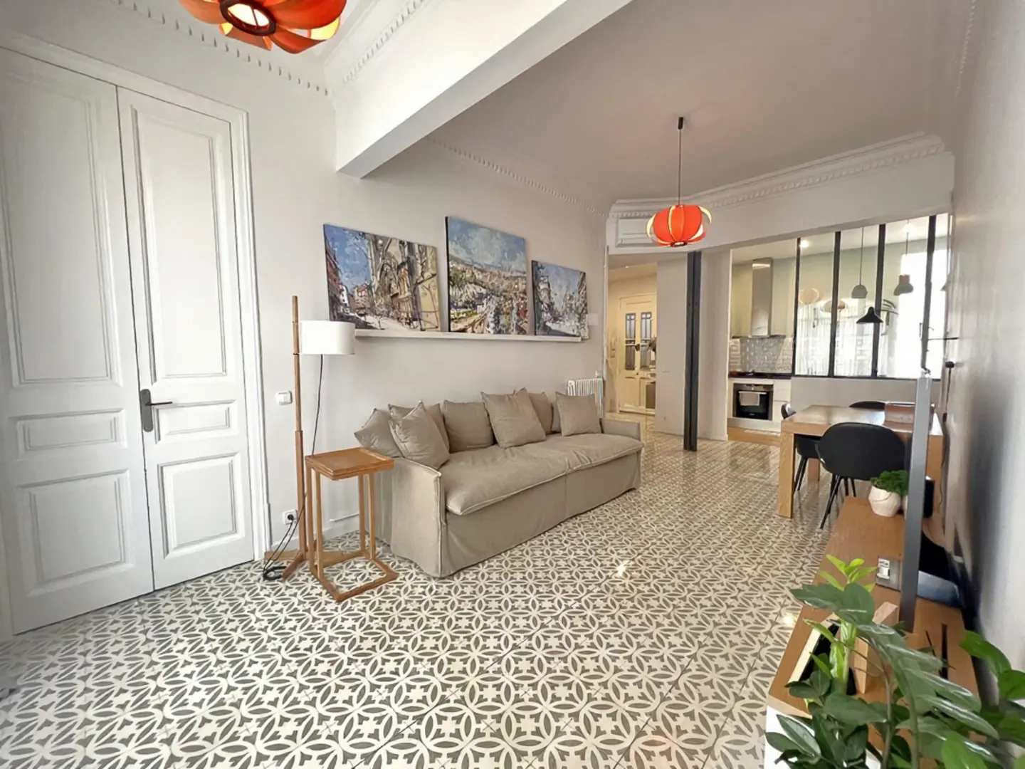 A bright living room with a beige sofa, artwork, and patterned tile flooring. A kitchen is visible in the background.
