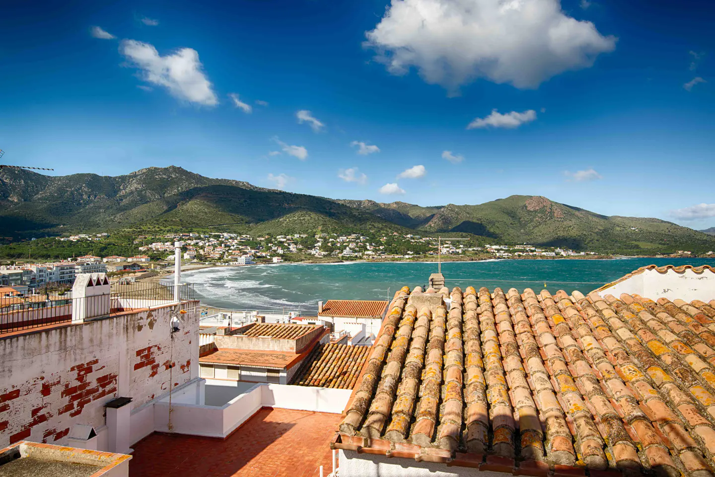 View from a rooftop with orange tiles overlooking a town by the sea, with mountains in the background under a blue sky with white clouds.