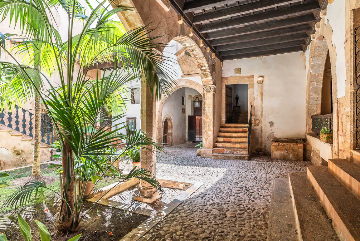 A sunlit stone courtyard with a palm tree, cobblestone floor, arched walkways, and a staircase leading to an open doorway.