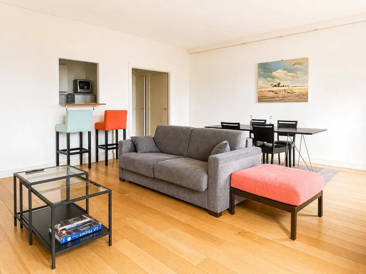Bright living room with a grey sofa, coral ottoman, glass coffee table, dining area, and colorful bar stools.