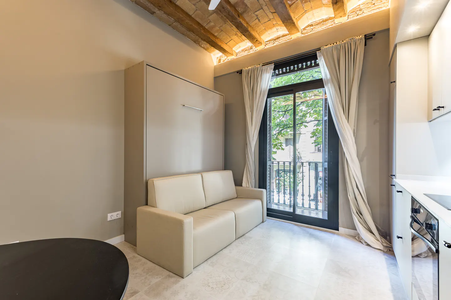 A modern studio apartment with a beige sofa, a Murphy bed, and a black-framed window with sheer curtains.