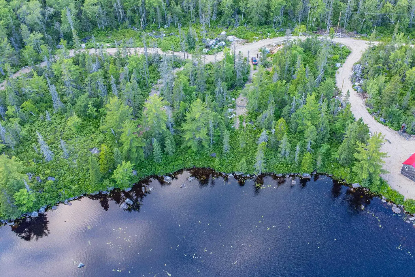 Aerial view of a dark blue lake surrounded by a green forest with a dirt road and a small red building.