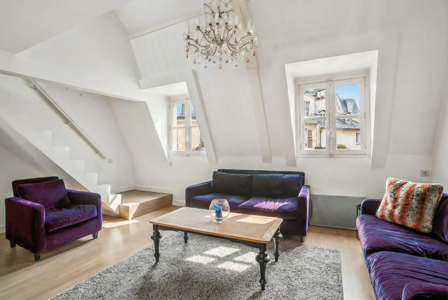 Bright attic living room with white walls, wood floors, and purple velvet furniture. A chandelier hangs above a coffee table on a gray rug. Windows show city views.
