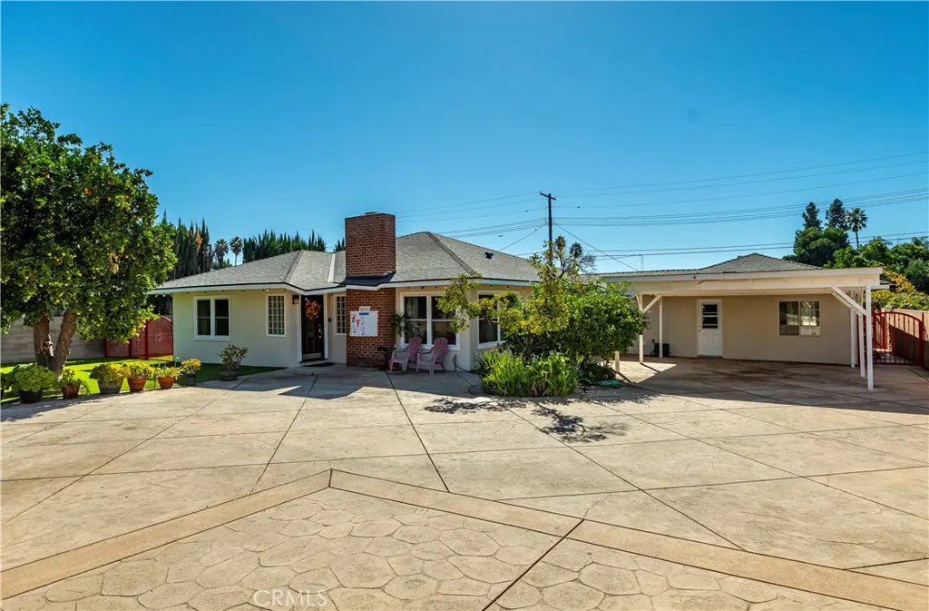 Beige single-story house with a brick chimney, a carport, and a large concrete driveway under a clear blue sky.
