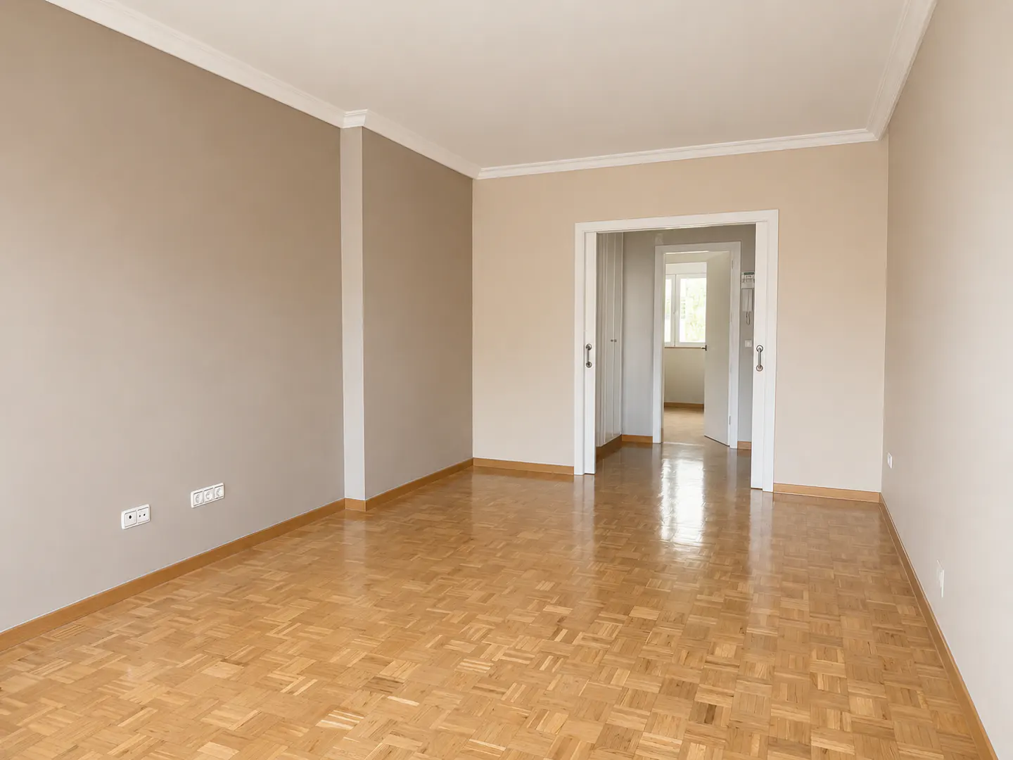 Empty room with beige walls, polished parquet wood flooring, and a doorway leading to a bright hallway.