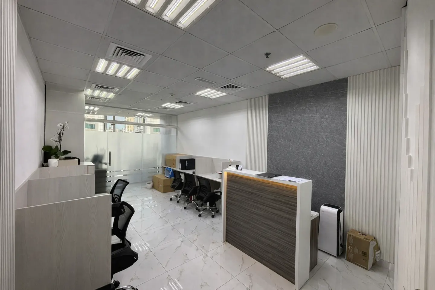 Modern office interior with a wood-paneled reception desk, white tiled floors, cubicle workstations, and frosted glass.