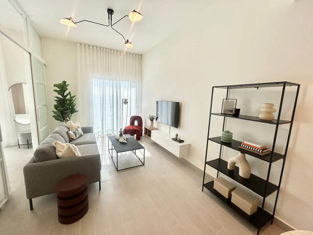 Bright living room with a gray sofa, black coffee table, and a black bookshelf against a white wall. A TV is mounted above a white console.