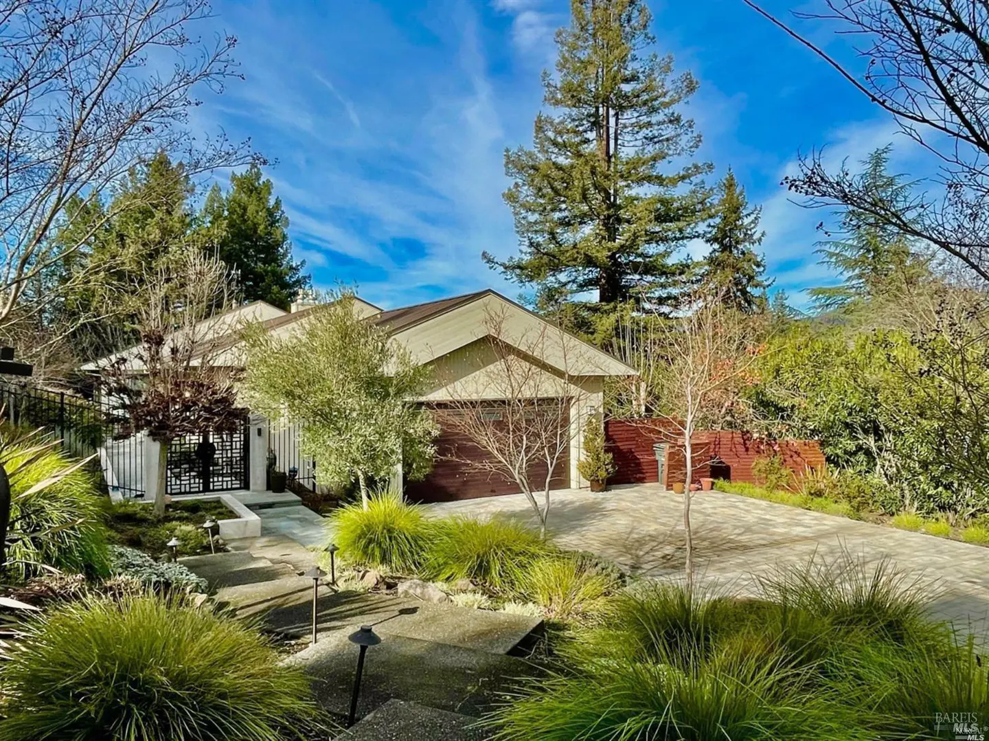 A tan house with a brown garage door is surrounded by green trees and bushes under a blue sky.