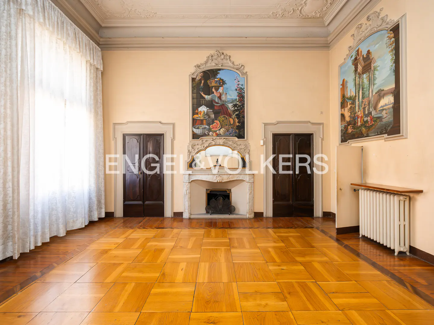 A spacious room with parquet floors, cream walls, and tall windows with white curtains. A fireplace with a painting above it is centered between two dark wood doors.