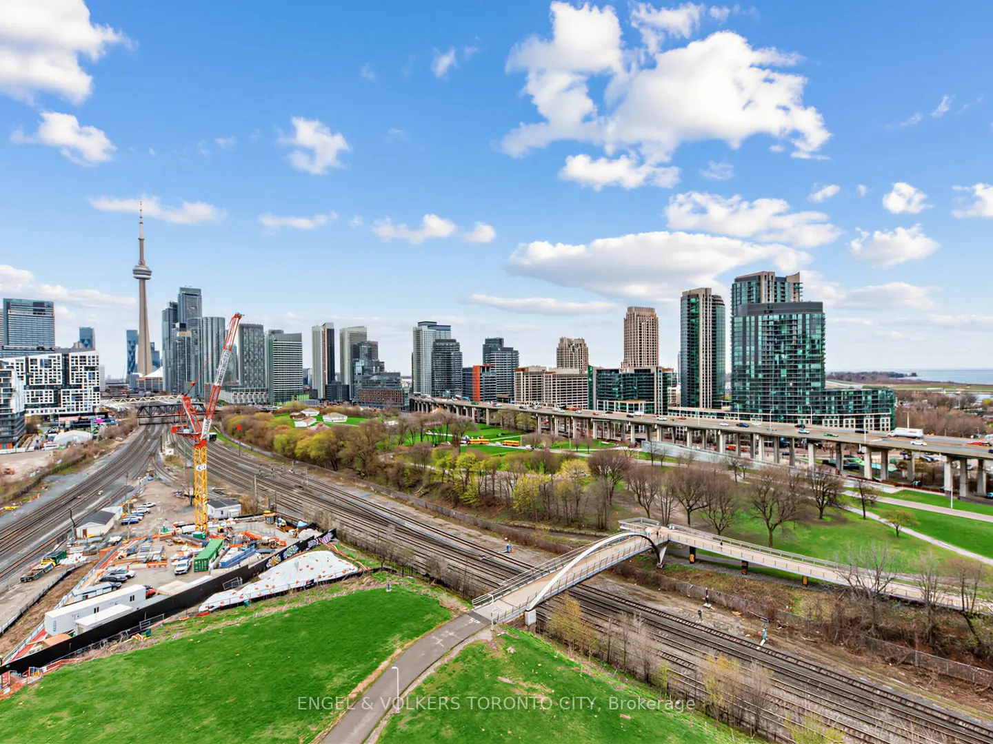 Aerial view of the Toronto skyline with the CN Tower, railway tracks, a pedestrian bridge, and green park space.